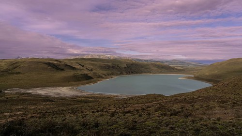 Turquoise lagoon in hilly steppe landscape at dusk, snow-capped Andes on the horizon under purple