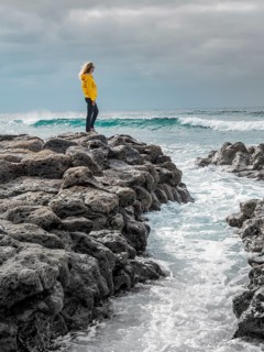 A woman in a yellow jacket stands on rugged volcanic rocks by the ocean, gazing at the waves under