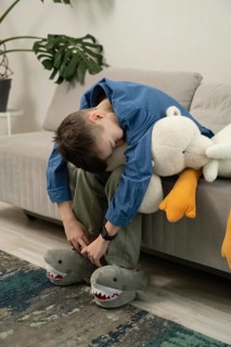 A boy in a blue shirt and green pants sits on a sofa cuddling a plush toy and wearing shark