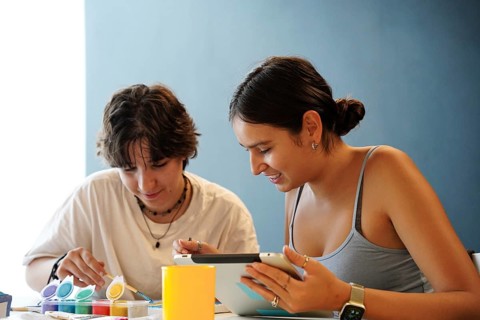 Two teenagers, one non-binary and the other female, are focused on painting and using a digital