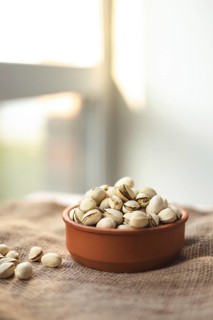 A rustic terracotta bowl filled with pistachios sits on a woven fabric. Sunlight streams in from