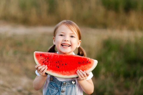 A joyful young child with pigtails holds a big slice of watermelon, smiling brightly during a warm