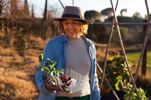 A woman in a hat and casual attire enjoys gardening outdoors, holding a plastic container with