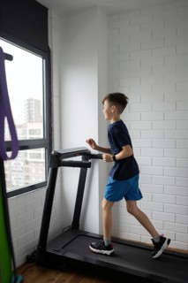 A young boy in a blue sports outfit is jogging on a treadmill inside a bright room with white brick