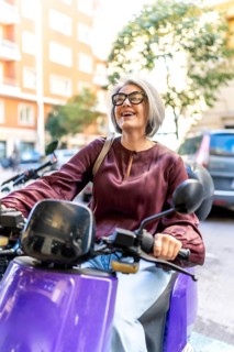 Senior woman with gray hair and glasses smiling brightly and looking up while riding a purple moped