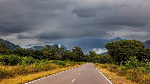 Lonely road leads through green countryside under a cloudy, stormy sky with mountains in the