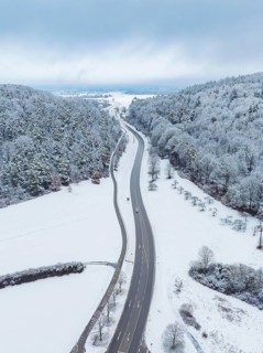 Snow-covered road through a wintry landscape with wooded hills and few moving cars, Aidlingen,