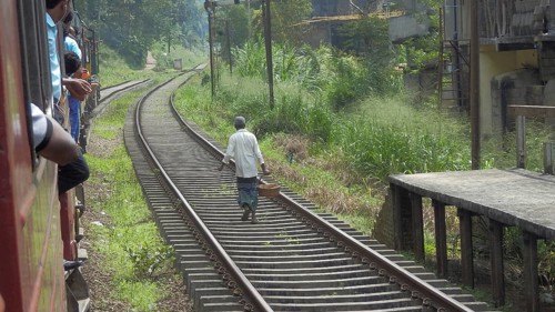 A man with a basket walks along the rails as passengers look out of a moving train, railroad