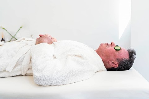 A mid-adult man enjoys a calming spa session. He lies down, eyes covered with cucumber slices,