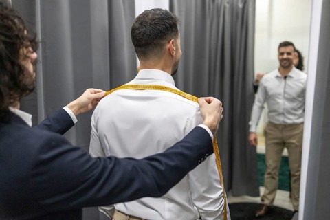 A professional tailor measures a client shoulder width using a tape measure in a fitting room. Both