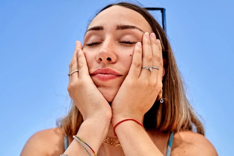 A woman enjoys a peaceful moment on the beach, with hands resting on their face, feeling the warmth