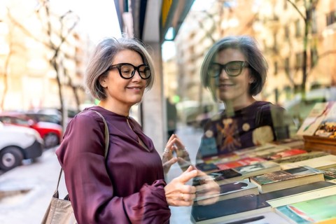 Mature woman with gray hair and eyeglasses smiling while carefully looking at a book display in a