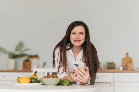 A nutritionist in a kitchen setting, holding a smartphone, surrounded by fresh produce. The