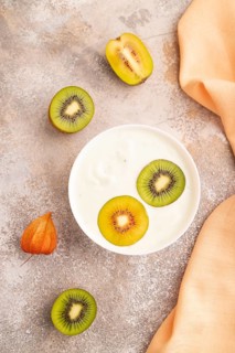 Yogurt with kiwi in white bowl on brown concrete background and orange linen textile, top view,