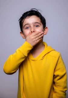 A young boy wearing a bright yellow hoodie covers his mouth with his hand, conveying surprise or