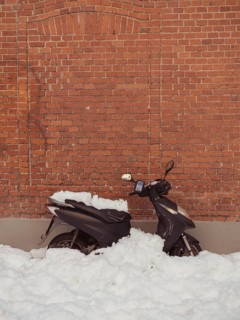 A lone scooter stands partially buried in thick snowdrifts against a weathered brick wall,