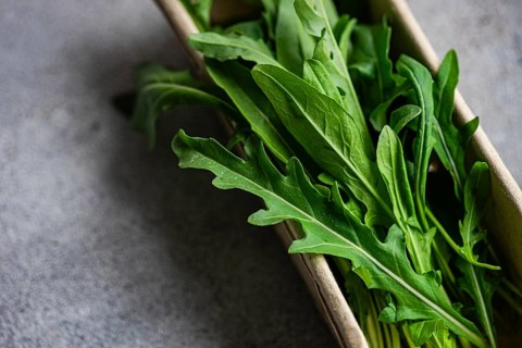 Close up of fresh, vibrant green arugula leaves stacked in a cardboard box. The leaves are crisp