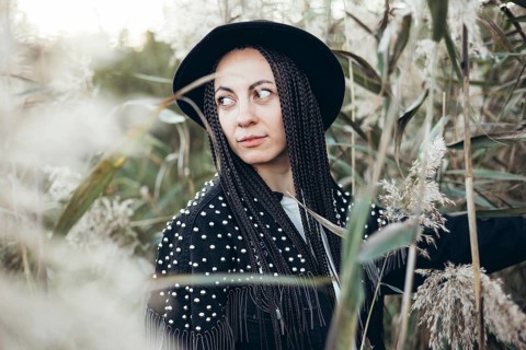 A woman stands amidst tall grasses at sunset, her braided hair gently catching the last light of