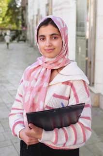 A arab student woman wearing a pink hijab, holding a folder while standing on a city sidewalk. The