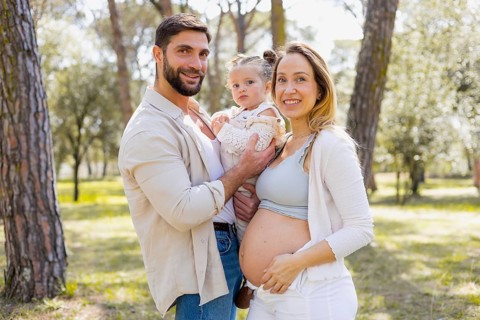 A pregnant woman stands with her partner and young child in a sunlit park. They are smiling and