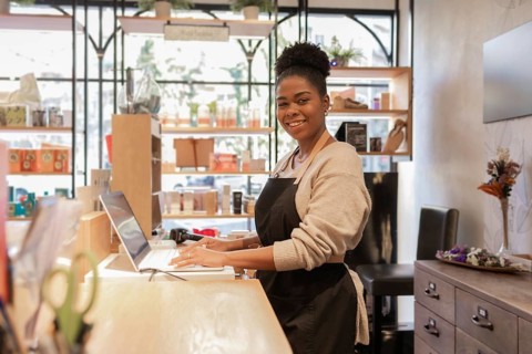 A cheerful store worker wearing a black apron smiles while assisting customers at a cosmetics