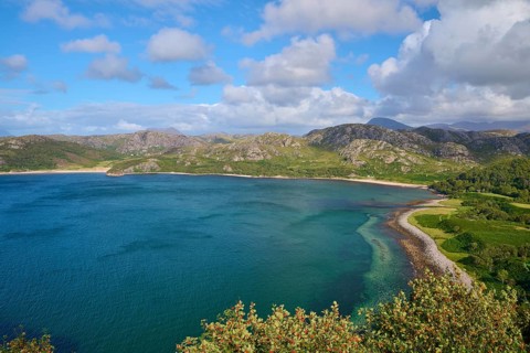 View of quiet bay surrounded by green mountains under a blue sky with white clouds, Gruinard Beach,