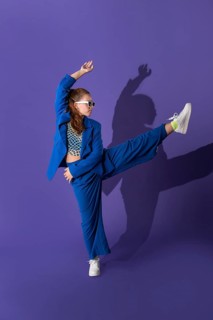 Young woman dancer striking a dynamic pose against a vibrant purple background. She wears a stylish