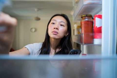 A woman stands in front of an open fridge, using her smartphone to shop for groceries online. The