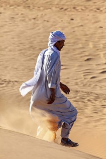 Man in traditional dress running through the desert, kicking up dust as he descends a sand dune, A