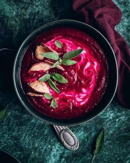 Overhead view of a beetroot soup garnished with slices of apple and fresh herbs. The rich, deep red