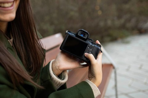 A woman in a green coat is smiling while holding a camera, ready to capture a moment The background