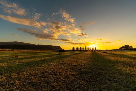 Moai statues of Ahu Tongariki as silhouettes at sunrise, grassy area in the foreground, cloudy sky,