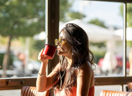 A woman with sunglasses sips from a red mug in a sunlit cafe. She enjoys a peaceful moment, seated