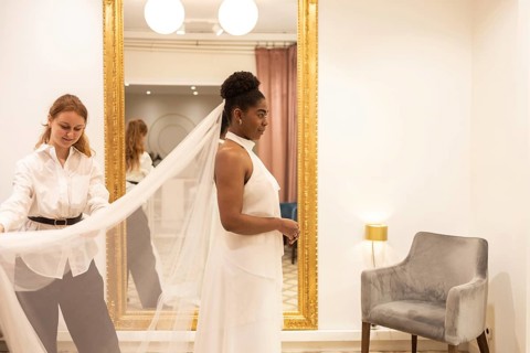 A bride stands in an elegant boutique as a stylist adjusts her veil. The scene captures the