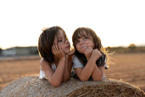 Two children lie on a hay bale in a sunlit field, enjoying each other's company. The warm golden