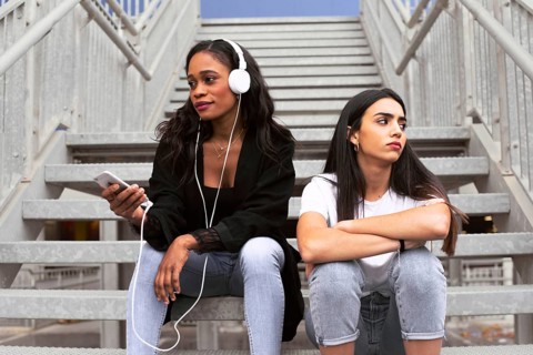 Two young women sit on a staircase. One listens to music with headphones, holding a smartphone,