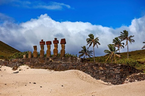 Moai statues on Easter Island against a partly cloudy, blue sky and surrounded by palm trees, The
