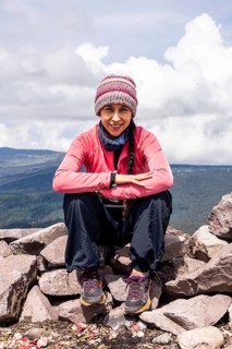 A smiling hiker in a colorful beanie and pink jacket takes a break on a rocky mountain top. The