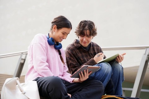A non-binary teen and a female student sit outdoors, engaging with digital tablets. They share a
