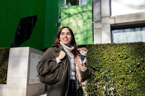A woman in a green jacket holds a small chihuahua while enjoying a sunny day outside a modern