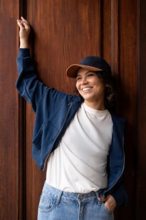 A woman smiles warmly, wearing a cap, blue jacket, white shirt, and jeans, standing against a