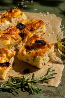 Golden homemade focaccia bread topped with rosemary and sun dried tomatoes, presented on parchment