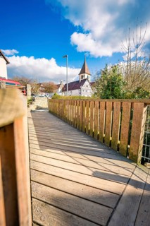 A wooden walkway leads to the village church, with bright skies and clear shadow lines, Deufringen,