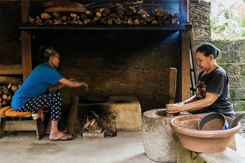 Two women engage in traditional cooking in a rustic kitchen. One uses a stone mortar for coffee