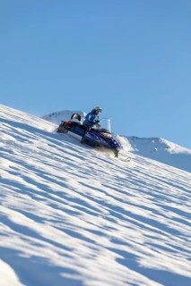 A person on a snowmobile speeds across a pristine, snowy slope with clear blue skies. Surrounded by