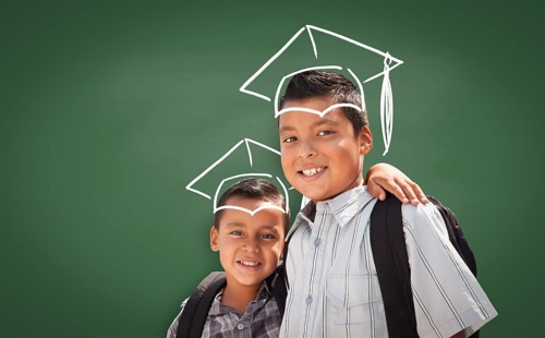 Young Hispanic Student Boys Wearing Backpacks In Front Of Blackboard with Graduation Caps Drawn In