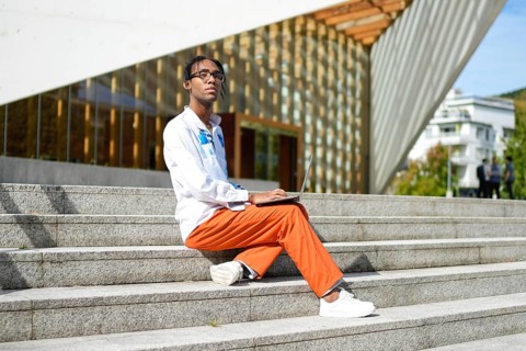 A man sits on outdoor steps with a laptop, embracing his gay pride. His reflective expression and