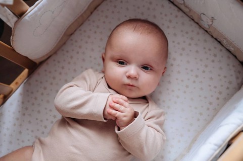 A baby boy lies comfortably in a crib, gazing thoughtfully. He wears a soft onesie, surrounded by a