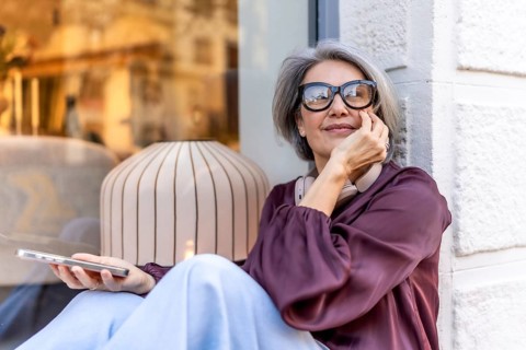 Grey haired woman sitting by a store window, holding her smartphone and wearing wireless headphones