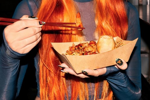 A woman with long orange hair is holding a paper tray of Asian street food, using chopsticks to eat